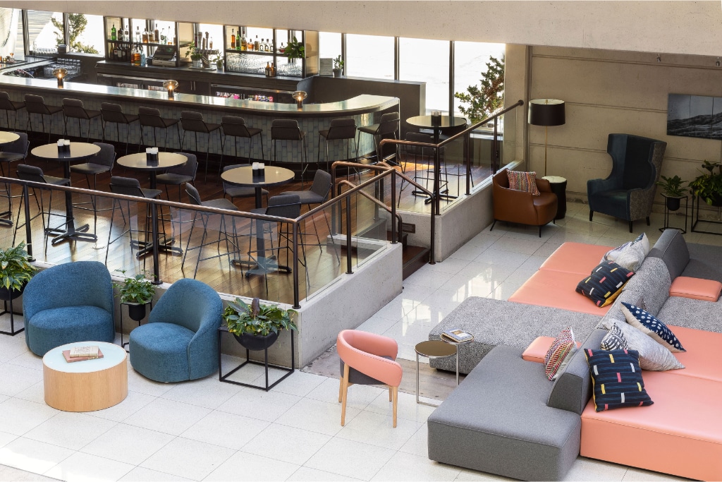 An open lobby area features a mix of seating options, including cozy chairs and a spacious sectional sofa. A bar with high stools is visible in the background, alongside decorative plants. Large windows allow natural light to illuminate the space.