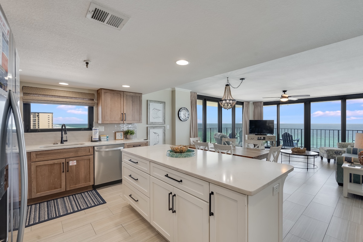 An open kitchen and living space is visible, featuring a large island with seating and a modern design. Natural light fills the area through expansive windows, showcasing views of the Gulf beyond. Kitchen appliances, including a refrigerator and dishwasher, are integrated into the cabinetry.