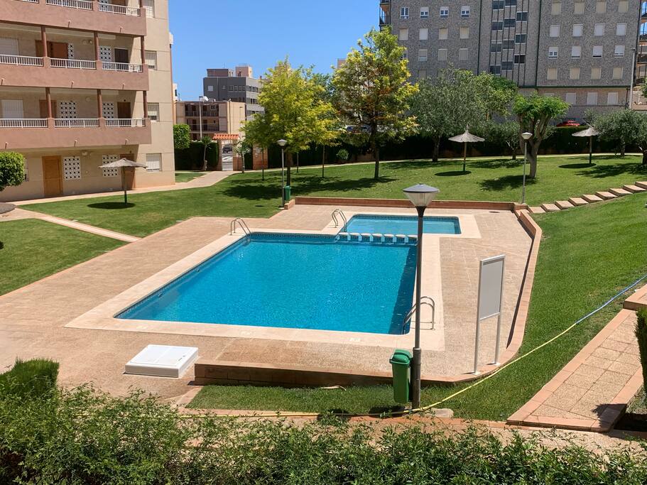 A well-maintained outdoor area features a clear swimming pool surrounded by lush greenery. Lounge chairs line the poolside, and shady umbrellas provide relaxation spots. The site includes manicured grass and pathways leading through the landscaped garden, creating a serene atmosphere.
