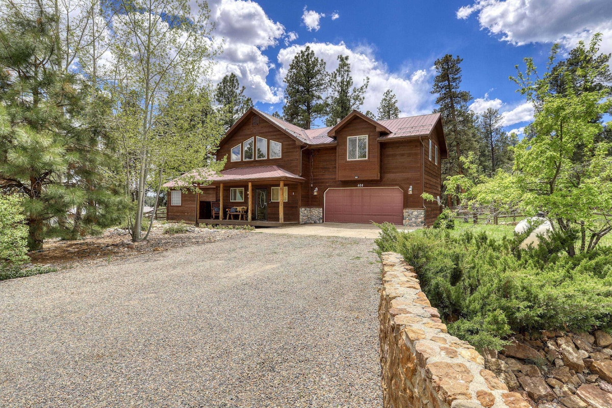 A two-story wooden house is set within a natural landscape, surrounded by tall trees and greenery. A spacious driveway leads up to the residence, which features a covered porch and large windows. The stone wall adds a rustic touch to the property's entrance.