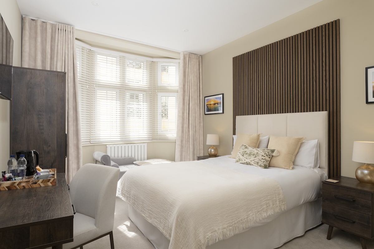 A well-appointed room features a spacious bed with light bedding and decorative pillows, accompanied by a wooden headboard. A desk with a chair is positioned near a large window, while soft curtains frame the space, allowing natural light to fill the room.