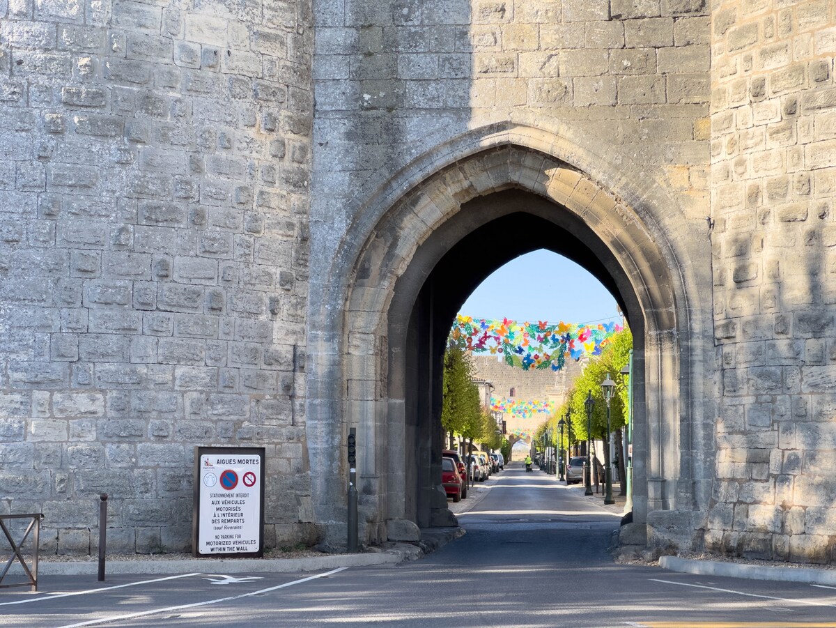 An archway made of stone leads into a street lined with trees. Colorful decorations hang across the entrance, adding a festive touch. A sign is positioned to the left, and parked cars are visible along the pathway.