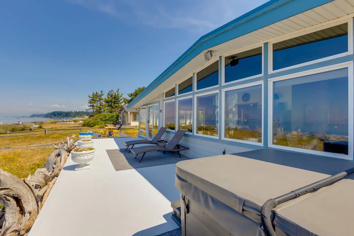 A spacious outdoor area is highlighted by a private hot tub and comfortable loungers positioned against a backdrop of large windows. The scenery includes a serene view of the coastline, with greenery and touches of sandy shore visible in the distance.