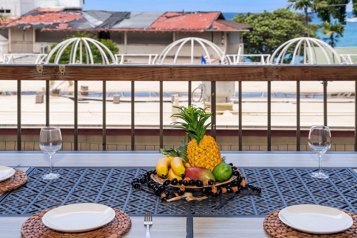 An outdoor dining table is set with four place settings, including glassware and woven placemats. A vibrant centerpiece featuring a pineapple and assorted fruits is displayed. The ocean and coastal scene with rooftops are visible in the background, framed by a railing.