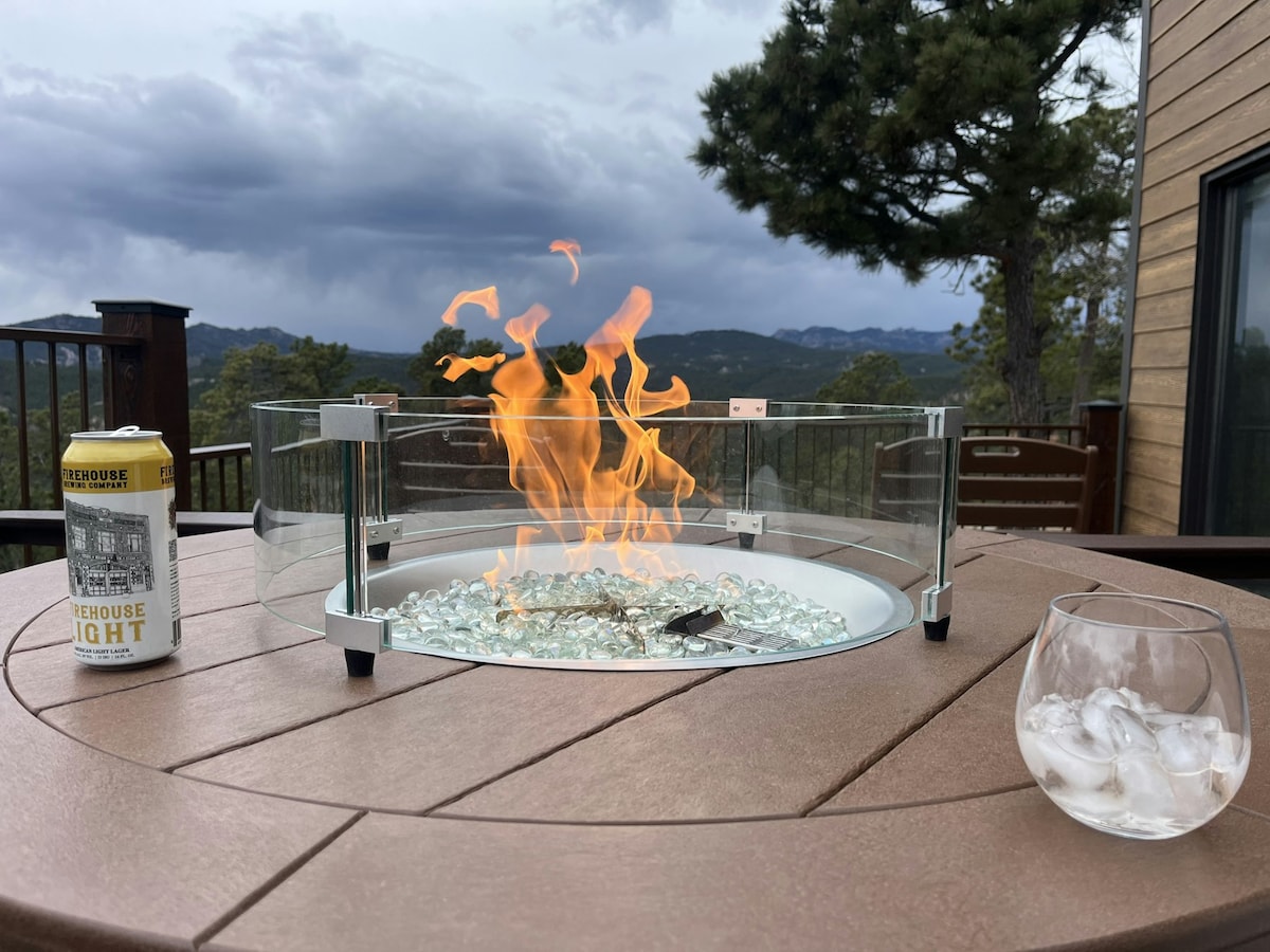 A propane fire table is featured on a wooden deck, surrounded by glass protection panels. Flames flicker above a bed of glass stones. Nearby, a glass filled with ice and a can of beverage are placed on the table, with a scenic mountainous backdrop under a cloudy sky.