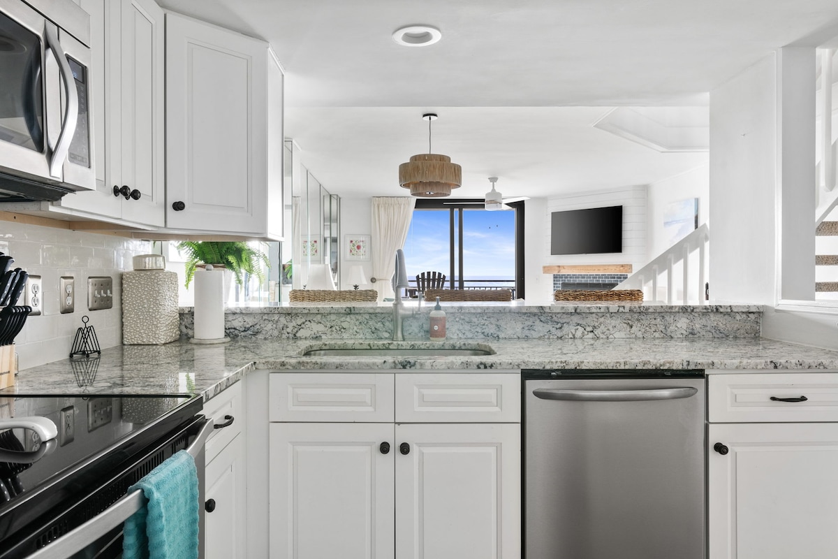 A modern kitchen is showcased with white cabinetry and a granite countertop. Stainless steel appliances are visible, including a microwave and dishwasher. A window offers a view of the ocean, while decorative items add a touch of style to the space.