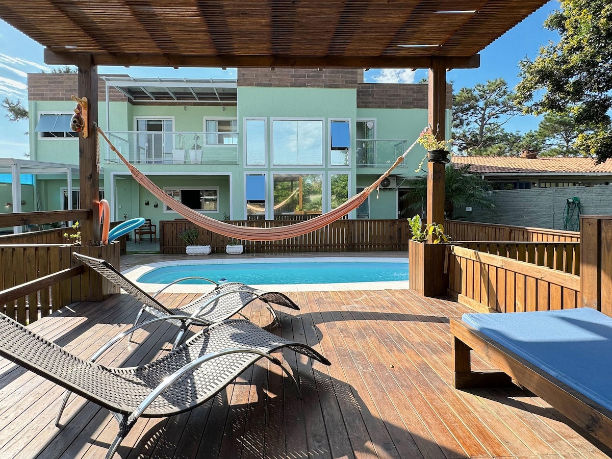 A wooden deck surrounds a refreshing pool, featuring lounging chairs and a hammock for relaxation. The background reveals a light green two-story house with large windows, allowing natural light to fill the space. Lush greenery enhances the tranquil atmosphere.