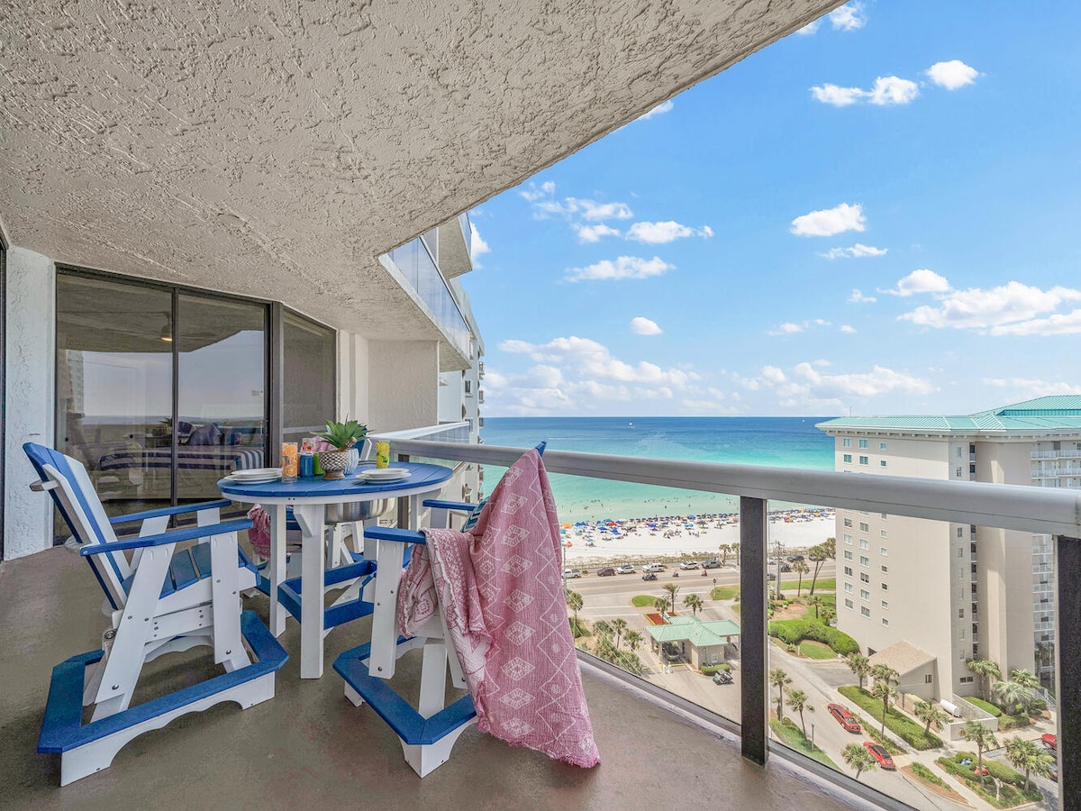 A balcony with blue and white outdoor furniture offers a dining area with a round table, surrounded by two adirondack chairs. A pink throw is draped over one chair, with sweeping views of the beach and Gulf waters visible in the background under a bright sky.