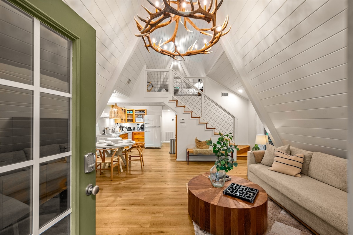 An open design living area invites relaxation, featuring a spacious sofa and a round coffee table. Above, an intricate antler chandelier draws attention, while a dining area with a wooden table is visible. Warm wood flooring complements the modern rustic decor.