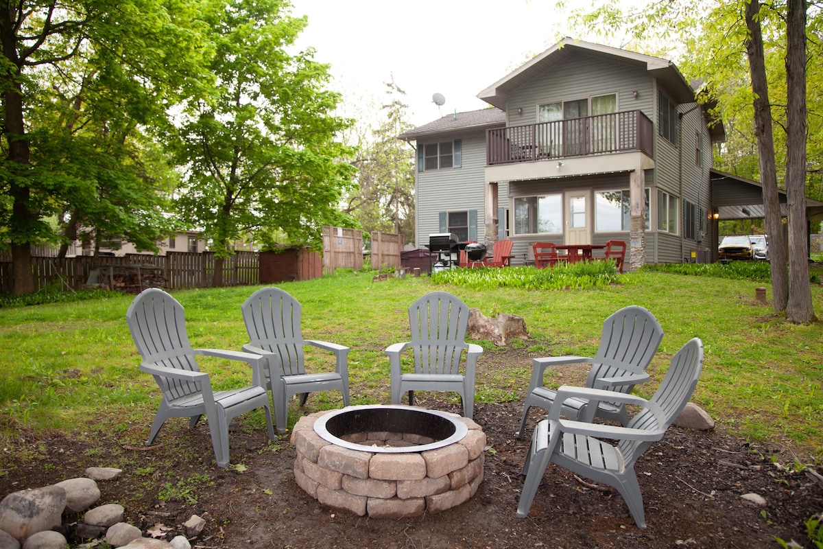 A spacious backyard features a circular stone fire pit surrounded by five gray Adirondack chairs. The well-maintained lawn is framed by trees, with a two-story house visible in the background, complete with balconies and outdoor seating.