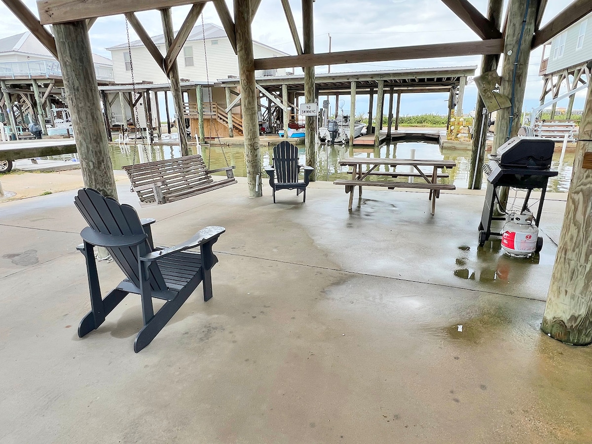 An open outdoor gathering area features a concrete floor with a gas grill and picnic table positioned under a wooden structure. Two Adirondack chairs and a swing provide seating options, with views of surrounding boat docks visible in the distance.
