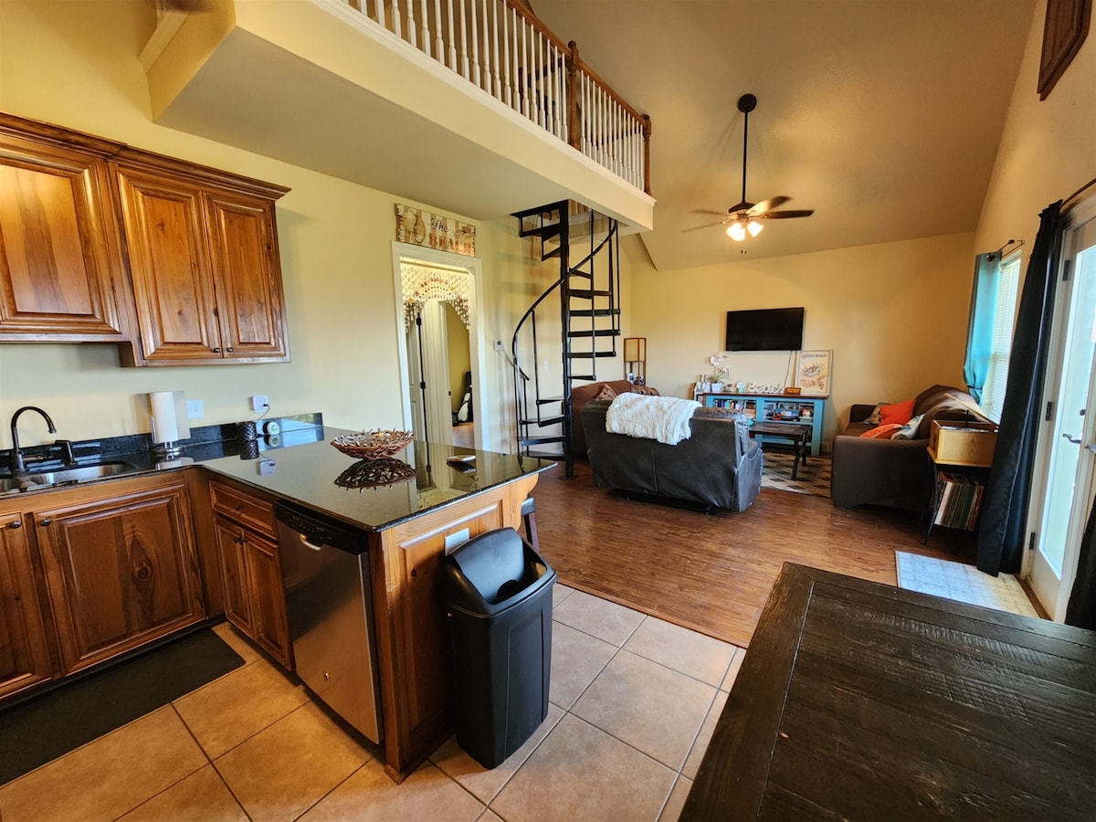 An open living area is visible, featuring a kitchen with wooden cabinetry and a black countertop. A dining area is set near a large window, while a spiral staircase leads to an upstairs loft. Natural light fills the space, creating a bright atmosphere.