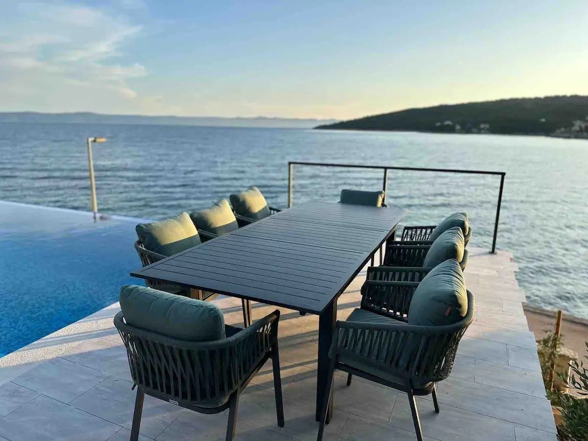 An outdoor dining area features a long table surrounded by six cushioned chairs, overlooking the coastline and the calm sea. A swimming pool is visible to the left, reflecting the soft colors of the sky during the evening light.