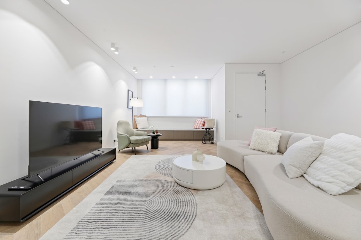 A modern living room features a curved sectional sofa, a round coffee table, and a large flat-screen TV on a sleek console. Soft lighting and natural sunlight filter through window treatments, creating a bright and airy space with minimalist decor.