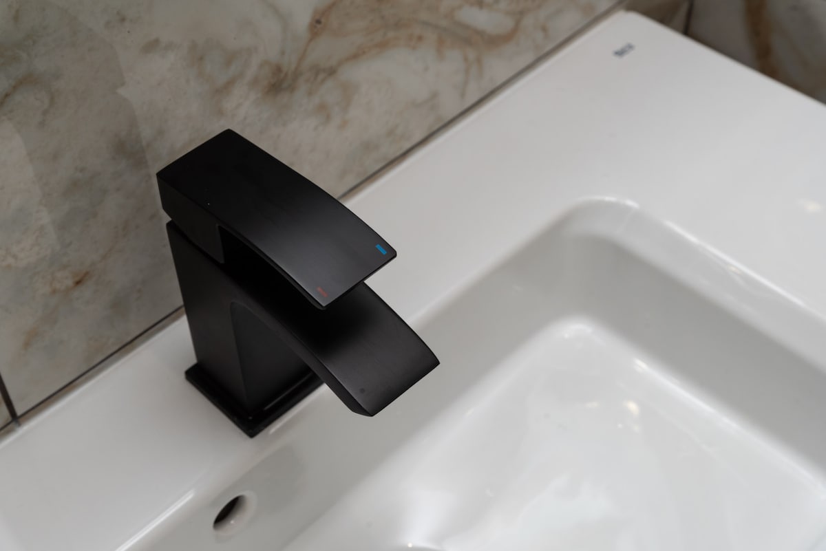 A stylish black faucet is positioned above a modern white sink. The sleek design features a minimalist spout with red and blue indicators for hot and cold water. The surrounding marble-effect tiles provide an elegant backdrop, enhancing the bathroom's luxurious feel.