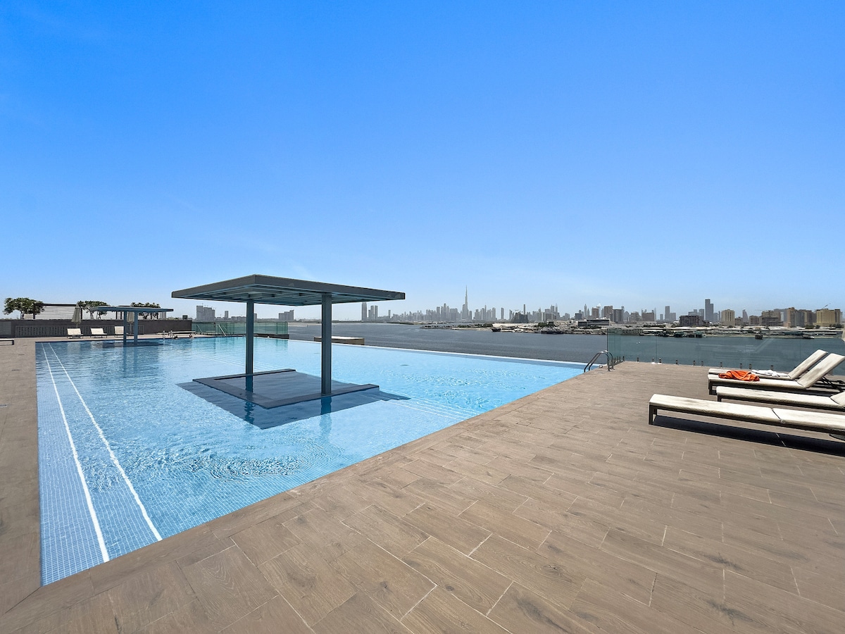 An infinity-edge swimming pool is set against a panoramic city skyline, with lounge chairs positioned along the deck. The serene water reflects the clear blue sky, creating a tranquil atmosphere. A shaded seating area is visible in the distance.