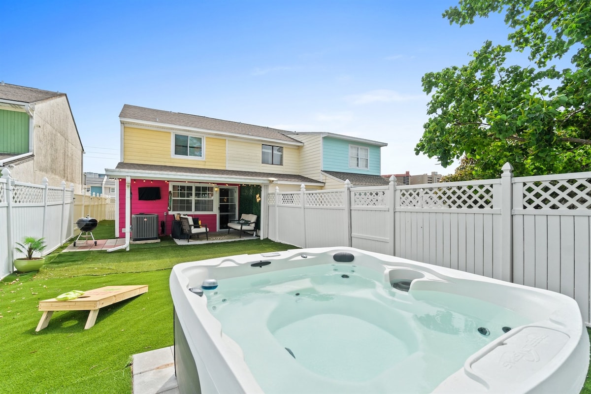 A private hot tub is situated in the backyard, surrounded by a well-maintained lawn and a white fence. The two-story house looms in the background, featuring a blend of pastel colors. A corn hole game area is visible on the grass, inviting outdoor leisure.