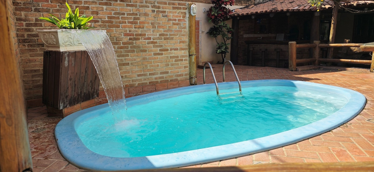 A pool surrounded by rustic brick walls features a gentle waterfall flowing into the clear blue water. The area is complemented by a potted plant, enhancing the natural elements present within the setting.