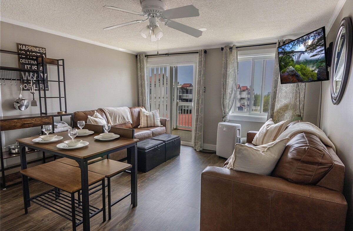 The living area features brown leather sofas and a sleek coffee table set for four. A 50" smart TV is mounted on the wall opposite the seating. Large windows allow natural light, showcasing the outdoor view. A ceiling fan is present for added comfort.