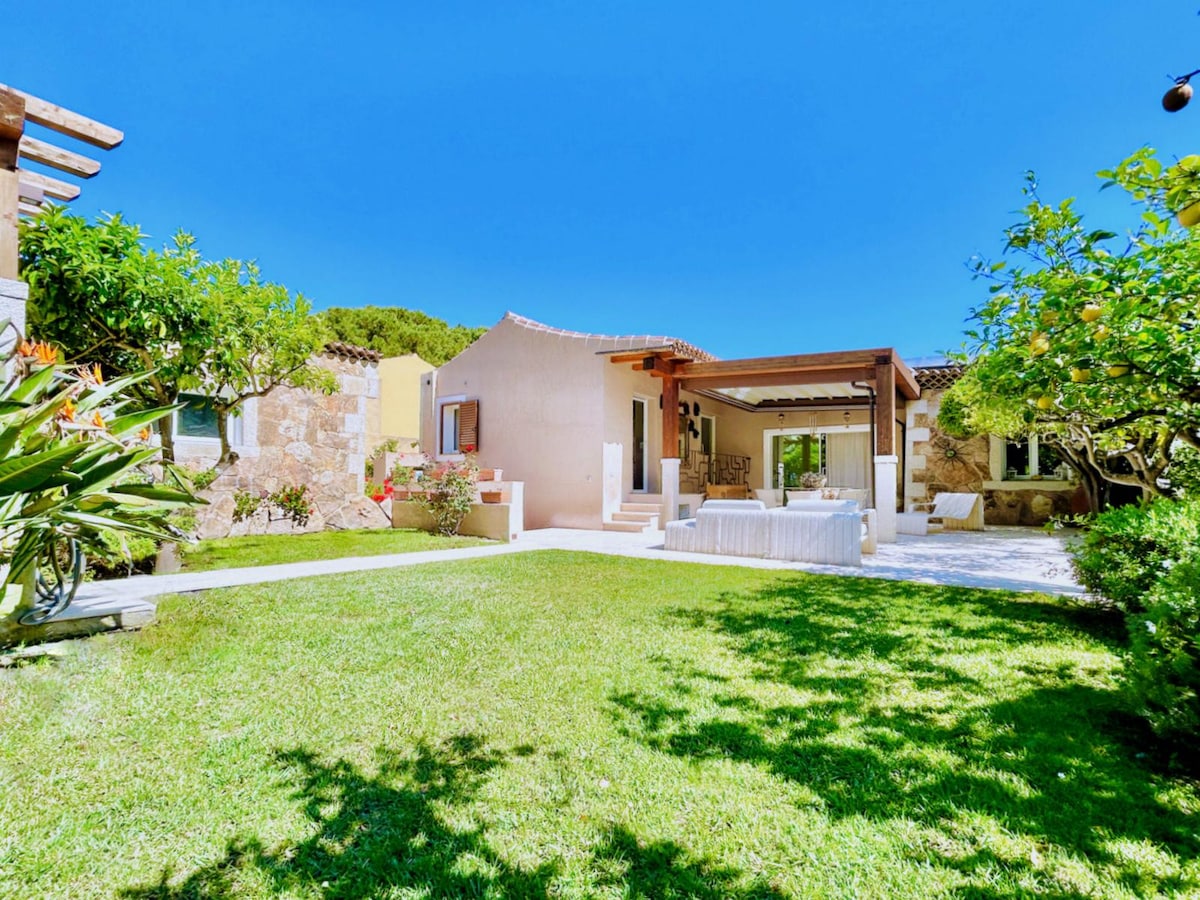 An outdoor view of a villa surrounded by lush green lawn and colorful plants. A covered veranda with seating is visible in the foreground, and the villa features a mix of stone and light-colored walls, blending harmoniously with the natural setting.