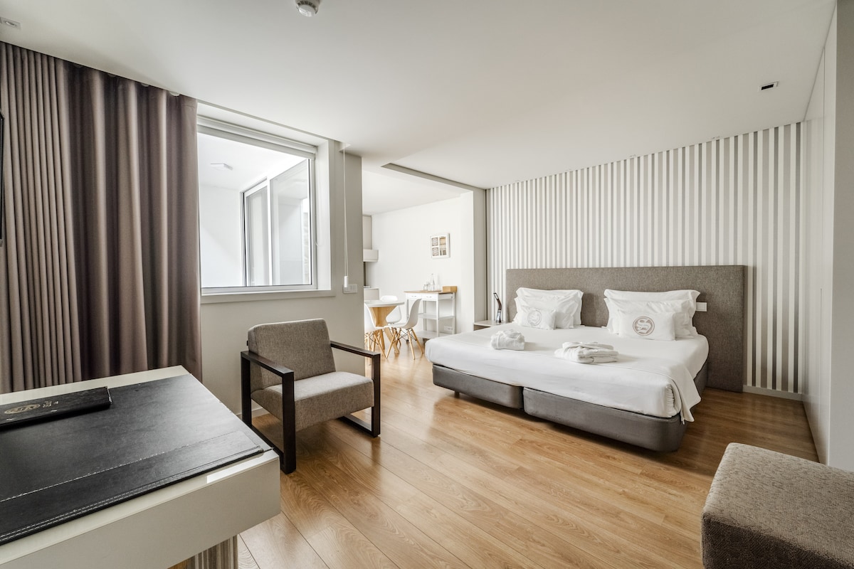 A bright room is seen with a spacious bed featuring neatly arranged linens. A large window allows natural light to fill the space. A small dining area with a table and chairs is visible in the background, contributing to the functional layout.