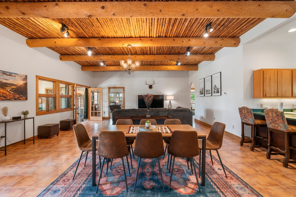 A spacious open living area is highlighted by exposed wooden beams and warm-toned flooring. A large dining table with brown chairs is centered, while a comfortable seating arrangement includes a dark sofa and a modern entertainment setup. Natural light fills the space through multiple windows.