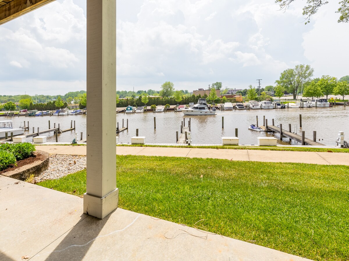 A view of the Black River is visible from the patio, showcasing docks lined with various watercraft. The grassy area leads to a concrete path, and a few trees frame the scene under a partly cloudy sky.