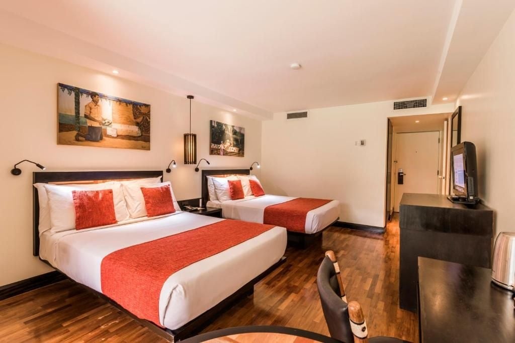 A well-lit guest room is presented, featuring two spacious beds with decorative orange accent pillows. Artwork adorns the walls, and a dark wooden desk with a chair is visible. Warm wood flooring adds to the room's inviting ambiance, and a flat-screen television is mounted on the wall.