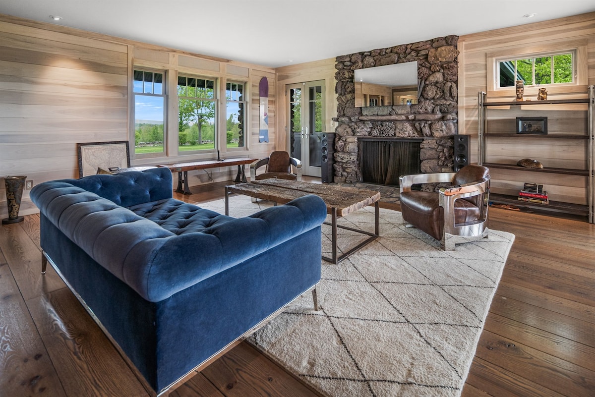 A spacious living area features large windows that invite natural light and offer views of the outdoor scenery. A deep blue tufted sofa is positioned alongside a wooden coffee table on a neutral area rug, complemented by two modern armchairs. A stone fireplace adds a rustic touch.