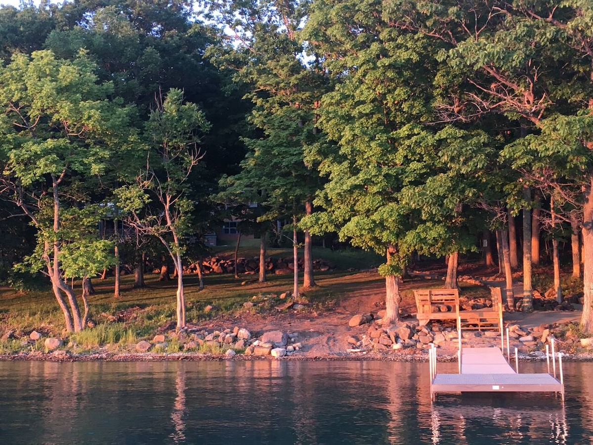 A serene lakeside view is captured, featuring a wooden dock at the water's edge. Lush green trees provide natural shade, while a grassy area is visible, dotted with stones. A bench offers a quiet spot to relax, surrounded by the calming presence of nature.