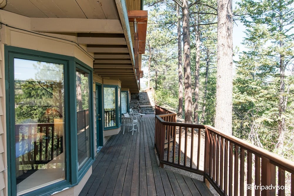 A spacious wooden deck extends alongside the lodge, framed by tall trees. Adjoining glass doors offer natural light, while outdoor seating is provided with white chairs and a table. The deck leads to a pathway, enhancing the connection with the surrounding nature.