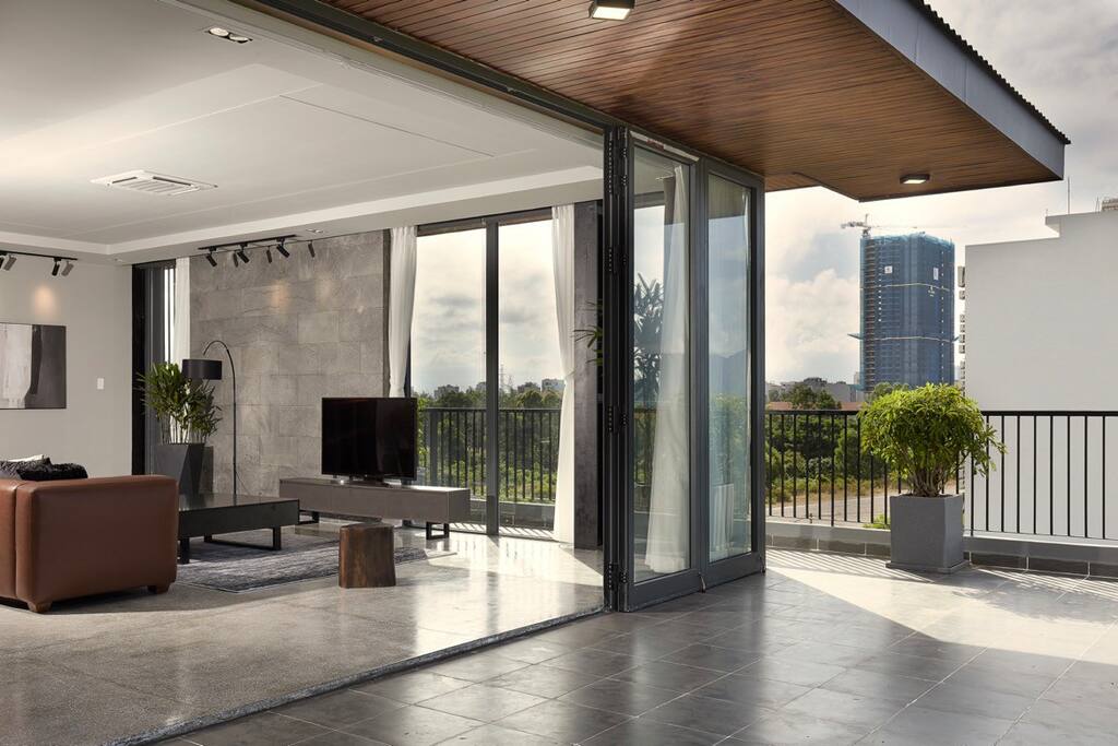 A spacious living room area features large floor-to-ceiling windows, allowing natural light to fill the space. The room is furnished with a modern sofa and a flat-screen TV, enhancing a sense of openness that extends to a private terrace.