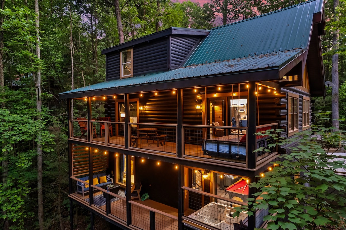 The cabin's exterior is framed by lush trees, featuring multiple levels with spacious decks illuminated by string lights. Comfortable seating areas are visible on each deck, inviting relaxation amidst nature. The metal roof complements the rustic wood siding, creating a harmonious blend with the surrounding forest.