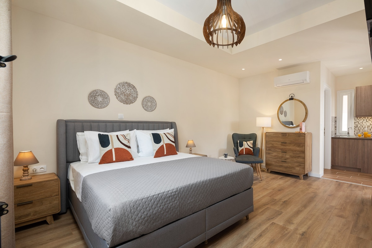 A spacious bedroom features a comfortable bed with decorative pillows and warm lighting from bedside lamps. A wooden dresser and a round mirror add a touch of elegance, while a cozy chair is placed nearby. The room is complemented by neutral tones and natural wood flooring.