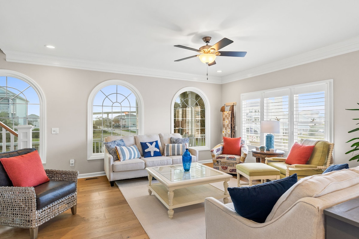 The living area features multiple windows with arched designs, allowing natural light to brighten the space. A mix of seating options, including a sofa and armchairs in various styles and colors, surrounds a central coffee table.