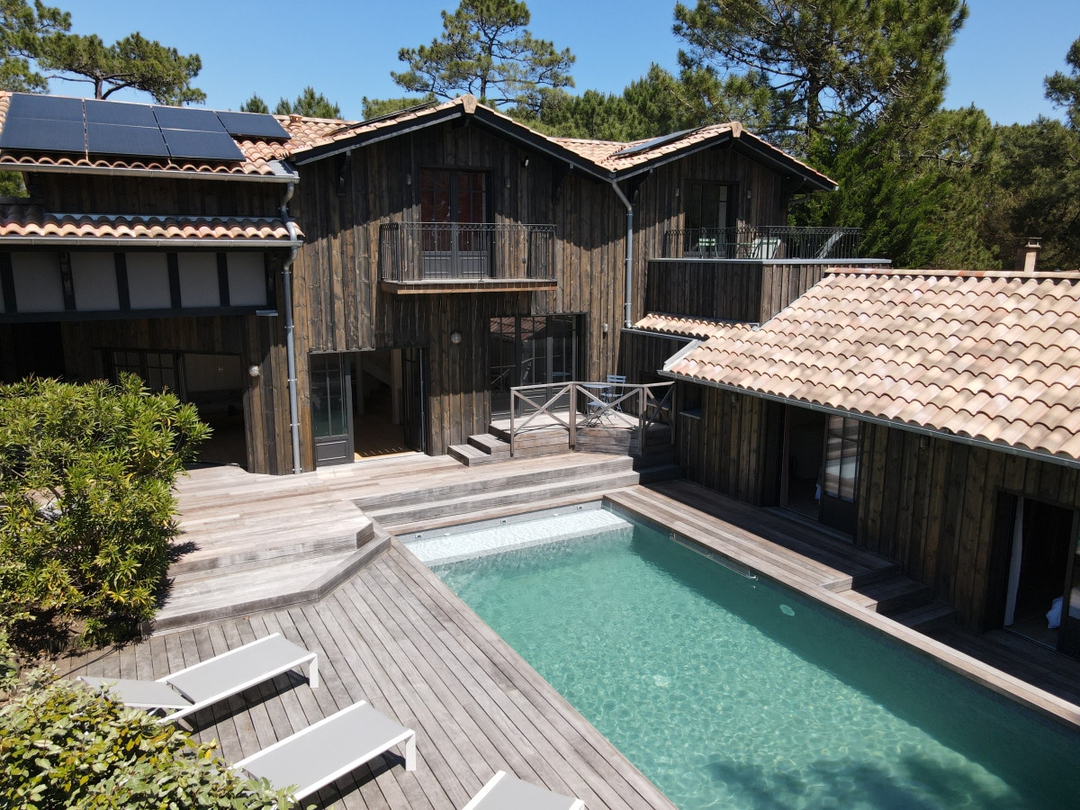 A modern wooden villa is surrounded by lush pines, featuring a private heated pool framed by wooden decking. Multiple levels and glass doors invite natural light, while sun loungers are arranged neatly beside the pool area. Solar panels are visible on the roof.