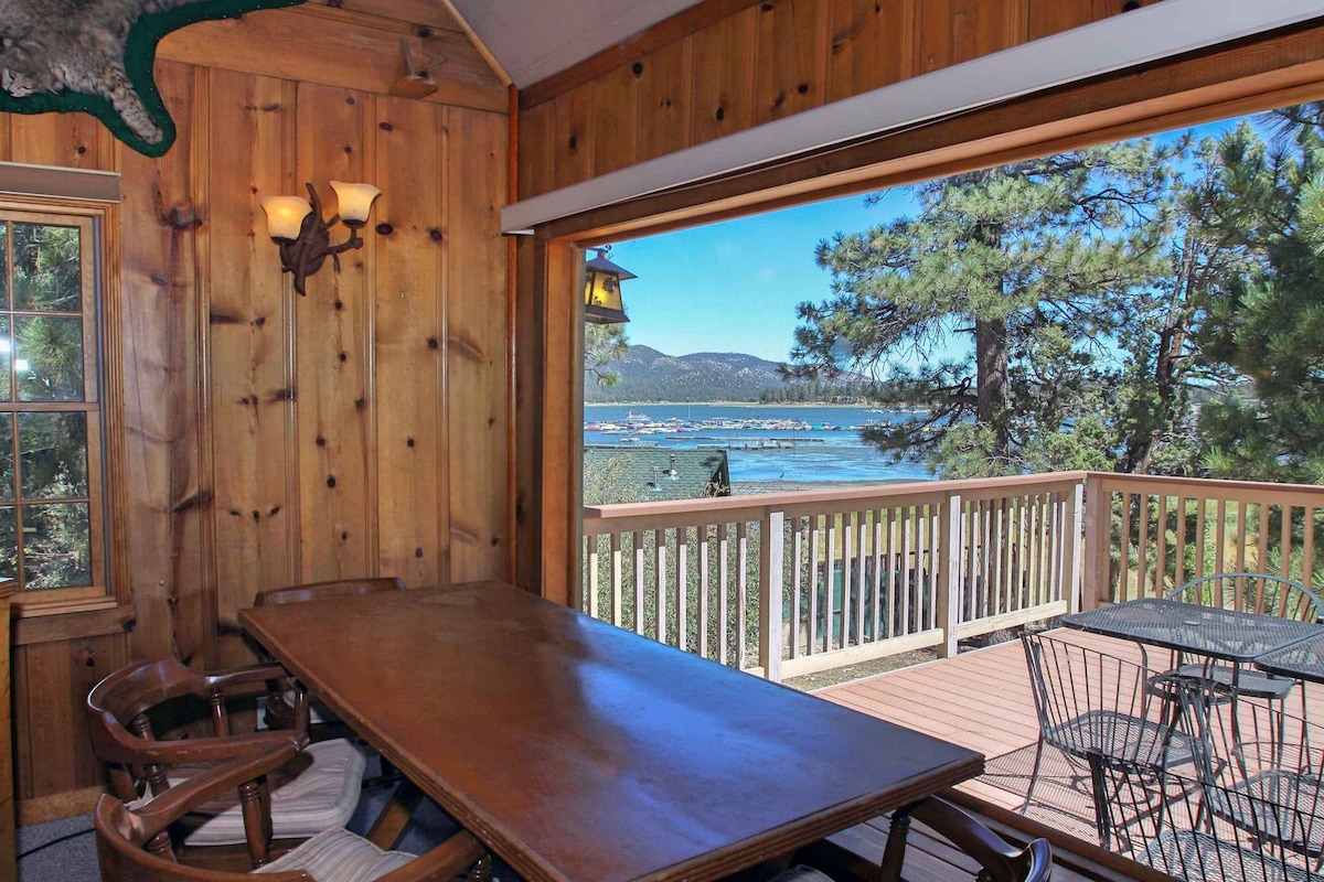 A dining area features a wooden table surrounded by four chairs, with a large window offering views of Big Bear Lake and the distant mountains. Natural light floods the space, highlighting the warm wood paneling and inviting atmosphere.