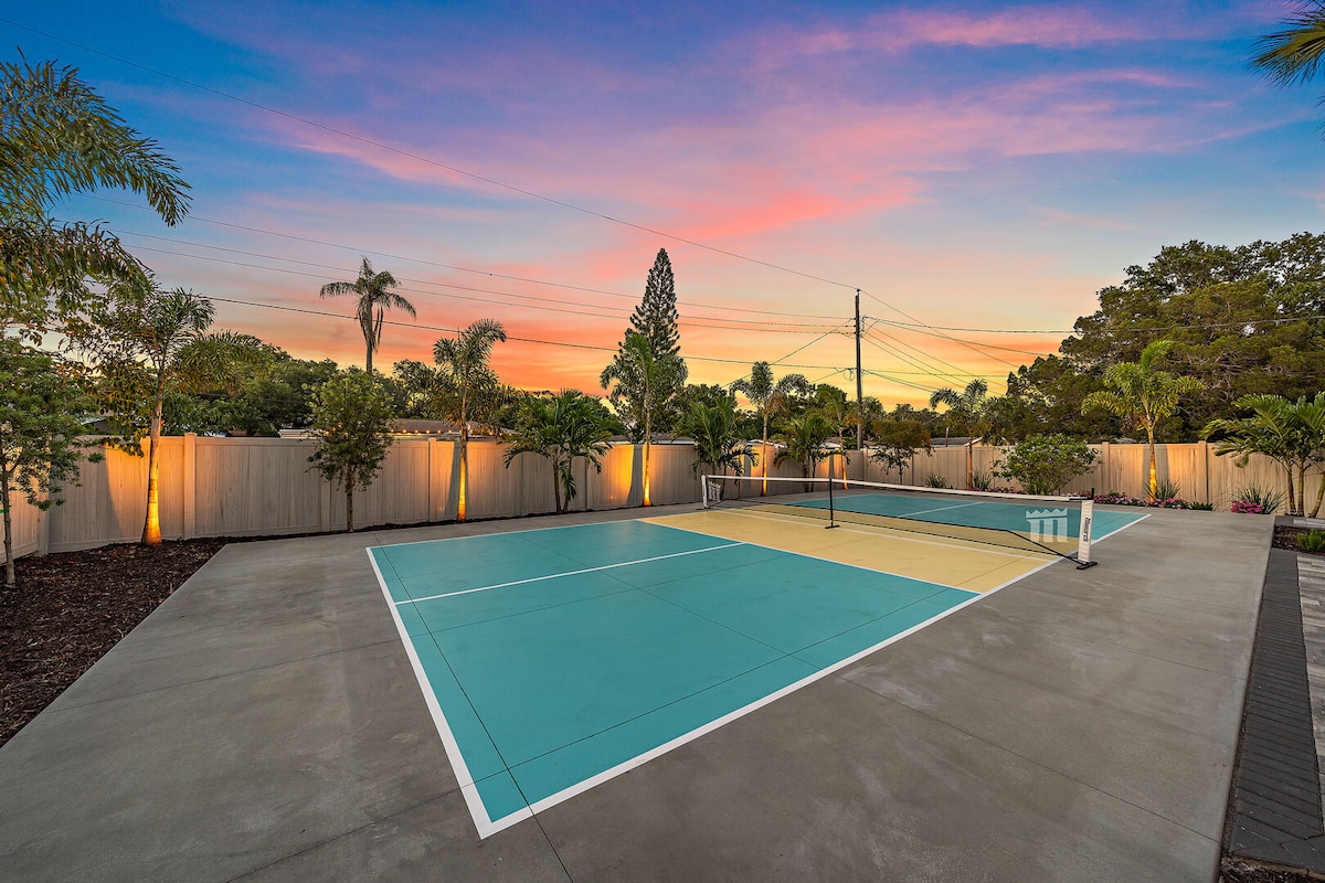 A regulation-size pickleball court is depicted, surrounded by lush greenery and a mix of palm trees. The court features vibrant turquoise and yellow surfaces, with soft lighting illuminating the perimeter. A colorful sunset sky enhances the serene atmosphere.