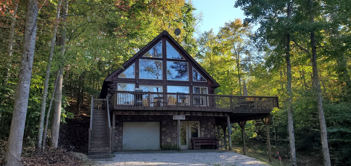 The cabin features a three-story design with large picture frame windows that offer a view of the surrounding trees. A spacious wrap-around deck is visible, providing access to the outdoor seating area. Stone accents create a natural aesthetic against the forest backdrop.