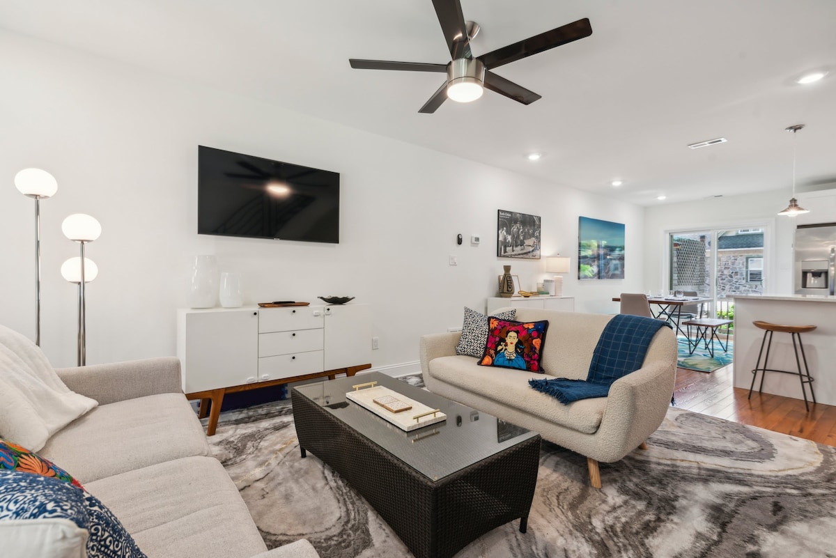 A spacious living area features a sleek, modern sofa and comfortable armchair, complemented by a decorative coffee table atop a stylish area rug. A flat-screen TV is mounted on the wall, and natural light fills the room from the adjoining kitchen and dining space.