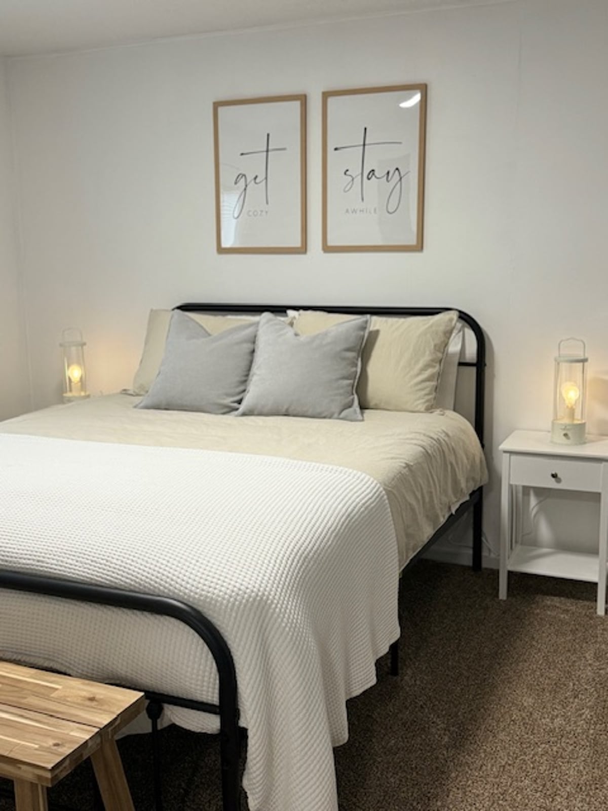 A well-lit sleeping area features a black metal bed with neutral linens and decorative pillows. Two bedside lanterns provide soft illumination. Above the bed, framed art with the words 'get' and 'stay' adds a welcoming touch to the simple decor.