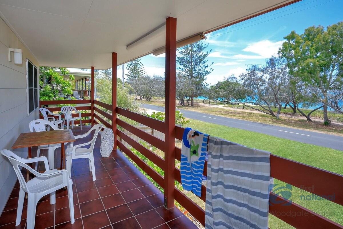 A covered outdoor patio area features a tiled floor and is equipped with white plastic chairs and a small table. Laundry items hang on a railing, with views of the green surroundings and glimpses of the ocean in the distance.