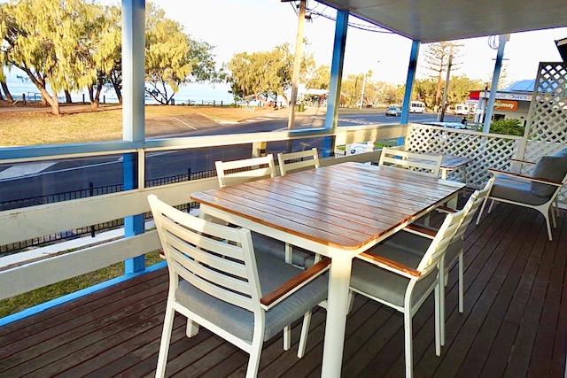 An outdoor patio area is furnished with a wooden dining table surrounded by six white chairs. The space is covered and features a wooden deck that offers views of the surrounding trees and road, with a glimpse of the coast in the distance.