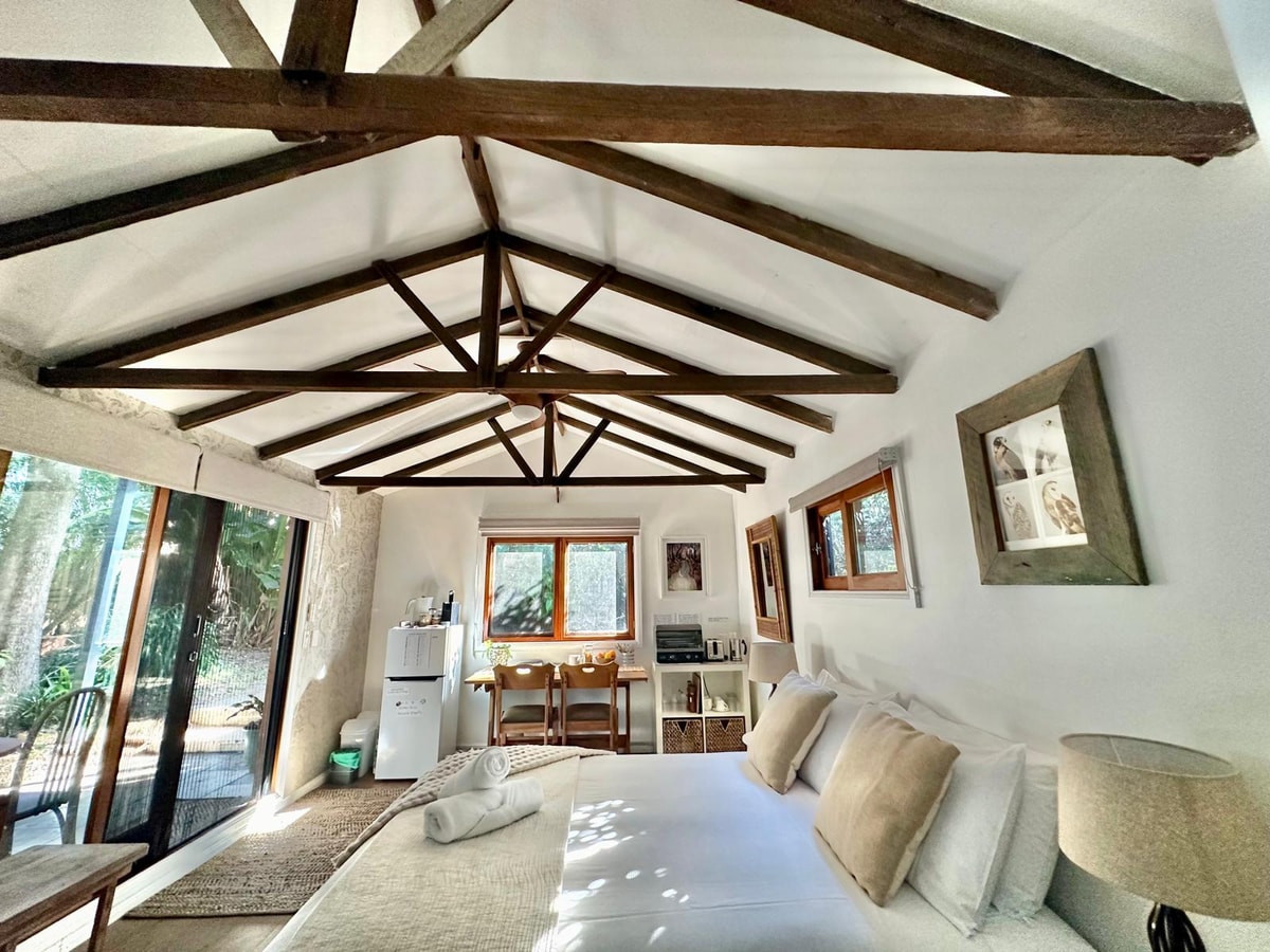 An open-concept living area features a spacious bed with neatly arranged pillows. Wooden beam ceilings enhance the airy feel. A kitchenette is visible with a refrigerator, while large windows provide natural light and a view of the surrounding greenery.