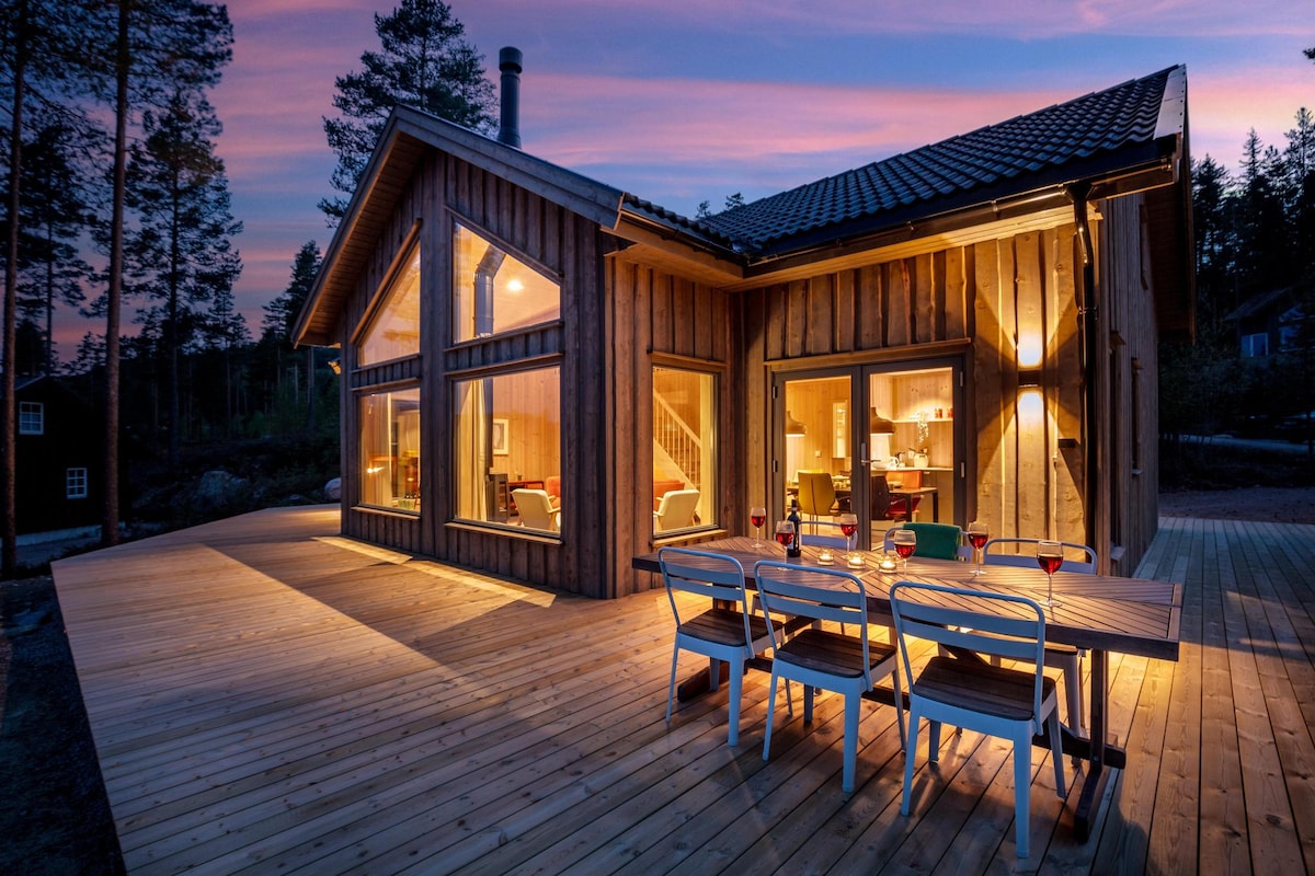 A wooden exterior of a modern vacation home is shown during sunset, with oversized windows reflecting warm light. A spacious deck features a dining area with multiple chairs and a table, inviting outdoor meals. The surrounding trees create a natural backdrop.