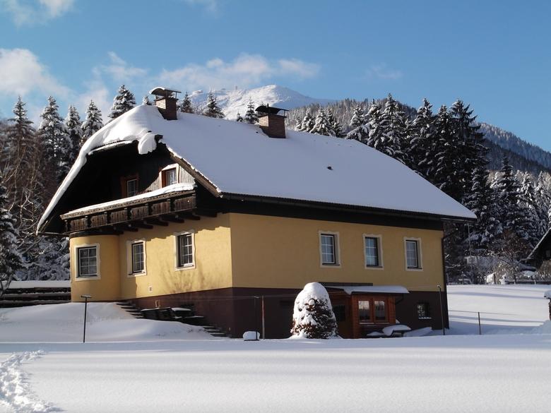 A charming two-story house is surrounded by snow-covered landscapes, with a sloped roof and overhanging eaves. The walls are painted in a warm yellow, contrasting with the white snow. Tall pine trees are visible in the background, enhancing the tranquil winter scene.