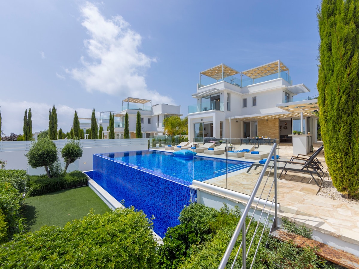 An expansive villa is showcased with a refreshing blue pool surrounded by sun loungers and shaded areas. Lush green landscaping complements the property. The architectural design features clean lines and multiple levels, inviting relaxation and enjoyment of the outdoor spaces under a clear blue sky.