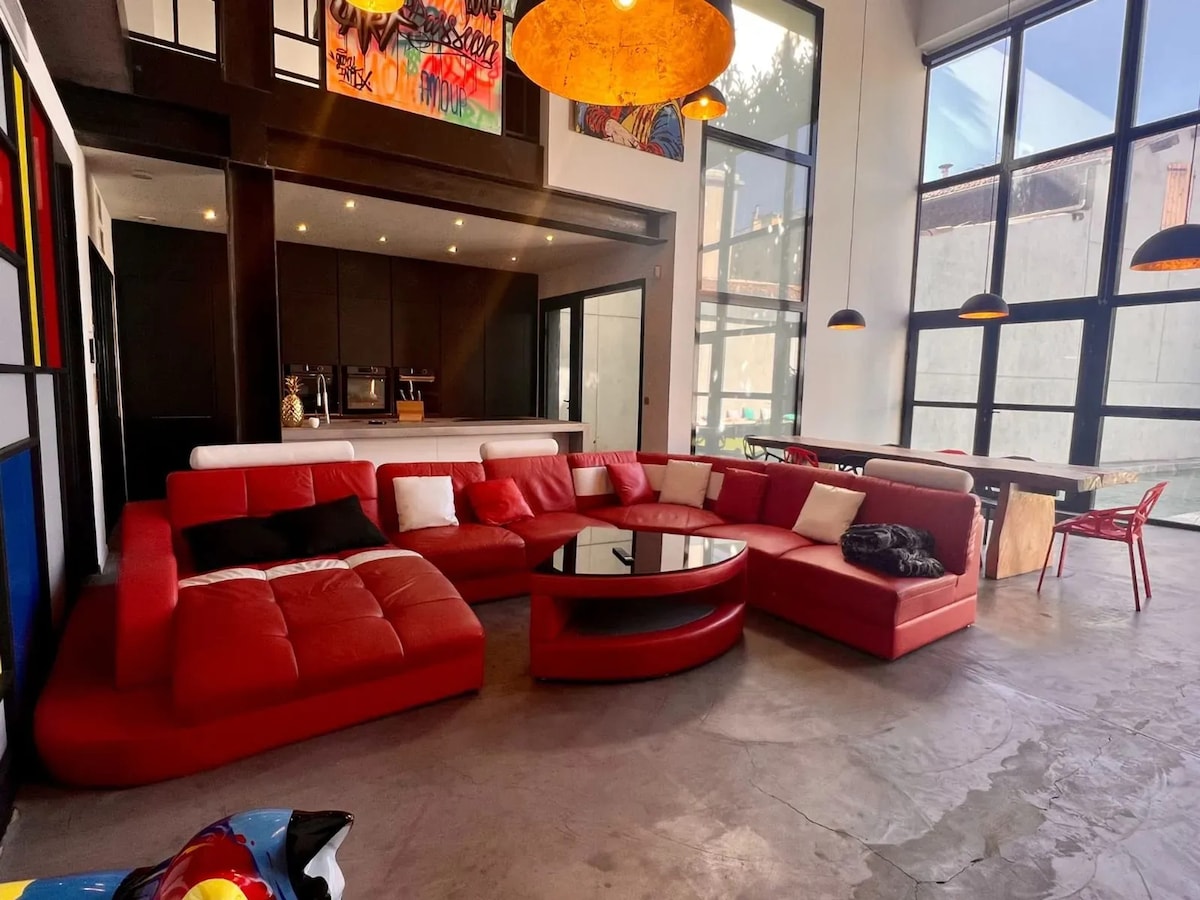 A spacious living area features a large red sectional sofa arranged around a circular coffee table. Bright artwork adorns the walls, and large windows allow natural light to fill the space. A visible dining area with modern chairs is included in the background.