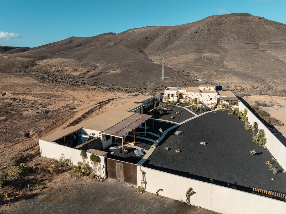 Aerial view of a rustic property enclosed by protective walls, surrounded by arid landscape. The structure features a large, open outdoor space with scattered vegetation and shaded areas. The distant mountains create a serene backdrop, enhancing the secluded atmosphere of the oasis.