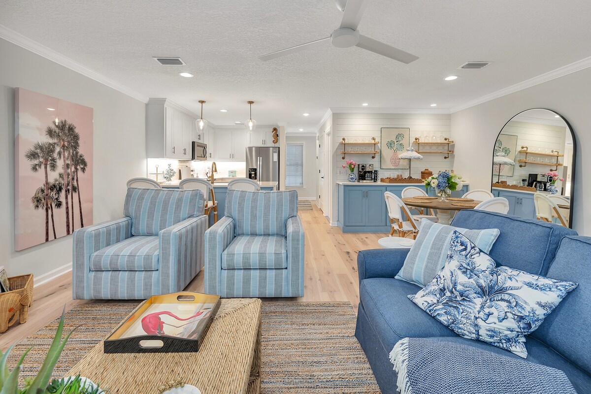 The living area features two striped chairs and a spacious blue sofa adorned with decorative pillows. A woven coffee table sits in the center, complemented by a textured area rug. The open layout leads to a bright dining area with a wooden table and well-equipped kitchen.